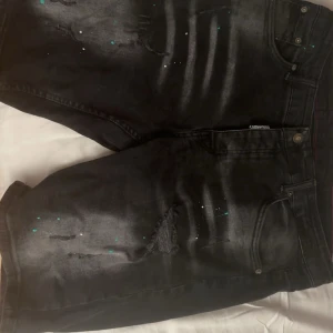 Svarta jeansshorts från Dsquared2 - Säljer ett par svarta jeansshorts från Dsquared2 med coola slitningar och färgstänk i turkos och vitt. Klassisk femficksmodell med knappgylf och Dsquared2-logga vid fickan. Perfekta för dig som gillar streetstyle och vill sticka ut lite extra.