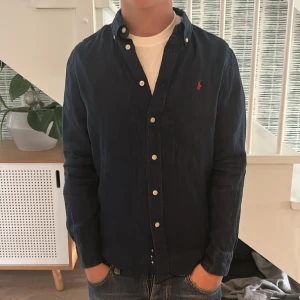  Ralph Lauren skjorta  - Snygg mörkblå skjorta från Ralph Lauren med klassisk krage och knappar hela vägen. Skjortan har lång ärm och ikonisk röd logga broderad på bröstet. Tillverkad i mjuk bomull som känns skön mot huden. Perfekt för dig som gillar stilrena och tidlösa plagg.