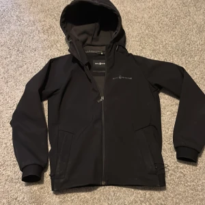 Svart vindjacka från Sail Racing - Svart vindjacka från Sail Racing med huva och hel dragkedja framtill. Jackan har två sidofickor med dragkedja och diskret logga på bröstet. Materialet är slätt och syntetiskt, perfekt för blåsiga dagar. Stilren och sportig look med normal passform. Nypris ca 1600kr. Pris är inte hugget i sten.