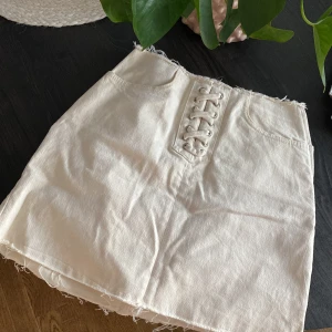 Vit kortkjol med snörning från NA-KD - Superfin vit kortkjol från NA-KD i jeansmaterial, storlek 36. Kjolen har snygg snörning framtill, råa kanter och dragkedja i sidan. Två fickor framtill ger extra stilpoäng.