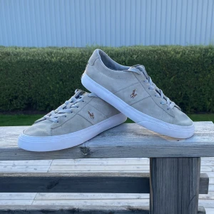 Beige mockasneakers från Polo Ralph Lauren - Snygga sneakers i beige mocka från Polo Ralph Lauren med klassisk logga broderad på sidan. Skorna har vit platt sula, rund tå och ljusgrå snörning. Perfekta för dig som gillar stilrena och tidlösa sneakers med premiumkänsla. 