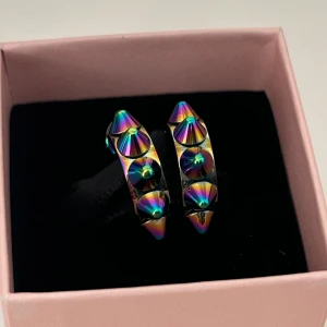 Edblad örhängen  - Coola örhängen i regnbågsfärgat metall med spetsiga nitar runt hela ringen. De har en chunky form och ger en edgy vibe till din stil. Perfekta för dig som vill sticka ut och addera färg och attityd till din look.