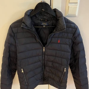 Mörkblå dunjacka från Polo Ralph Lauren - Snygg mörkblå dunjacka från Polo Ralph Lauren med klassisk röd logga på bröstet. Jackan har quiltad design, hög krage och två dragkedjeförsedda fickor. Perfekt för kyliga dagar och enkel att matcha med olika outfits. Jackan sitter perfekt på mig som är 175, inga defekter eller tecken på användning heller.