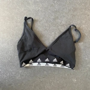 Svart linnetopp från Adidas - Svart linnetopp från Adidas med smala justerbara axelband och v-ringning. Resårbandet nedtill har Adidas-loggor i vitt runt om. Perfekt för träning eller chill dagar. Mjuk och skön bomullskänsla.