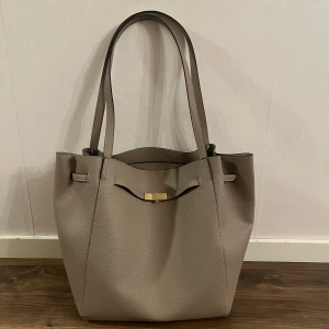 Beige axelväska från H&M i syntet - Stilren beige axelväska från H&M med guldigt spänne och långa handtag. Väskan är tillverkad i strukturerad syntet med polyester och har ett rymligt huvudfack samt innerficka med dragkedja. Perfekt för dig som vill ha en enkel men snygg väska till vardagen.