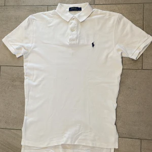 Vit pikétröja från Polo Ralph Lauren - Klassisk vit pikétröja från Polo Ralph Lauren med korta ärmar, krage och knäppning framtill. Broderad marinblå logga på bröstet. Tillverkad i mjuk bomull som känns skön mot huden. Perfekt för en clean och stilren look.
