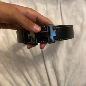 Svart hermesbälte  - Brett svart bälte i skinn med ett stort, blankt H-format metallspänne. Bältet har vita kontrastsömmar längs kanterna som ger en snygg detalj. Perfekt accessoar för att lyfta din outfit med en stilren och modern touch.