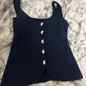 Mörkblå väst med silvriga knappar - Snygg mörkblå väst med breda axelband och unika silvriga knappar framtill. Västen har en figurnära passform och lätt rundad nederkant. Perfekt att styla över en skjorta eller t-shirt för en trendig look.