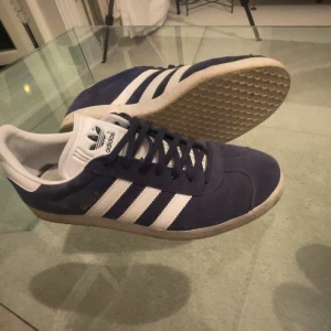 Adidas Gazelle sneakers mörkblå/vita - Säljer ett par klassiska Adidas Gazelle sneakers i mörkblå mocka med vita detaljer och de ikoniska tre ränderna på sidorna. Skorna har vit plös och häl, samt gummisula med mönster. Perfekta för dig som gillar retrostil och streetwear. Skick 7/10