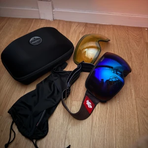 Blå Majesty skidglasögon - Säljer ett par snygga skidglasögon från Majesty med blå spegelglas och extra gul lins. Glasögonen har bred svart rem med röd logga och kommer med både skyddspåse och hårt fodral. Perfekt för dig som vill ha cool stil i backen.