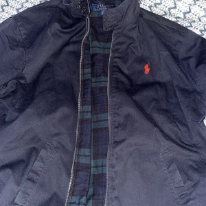Svart vindjacka från Polo Ralph Lauren - Mörkblå vindjacka från Polo Ralph Lauren med röd broderad logga på bröstet. Jackan har ståkrage med knappar, dragkedja framtill och två snedställda fickor. I princip helt ny, perfekt för dig som gillar klassisk och stilren look.
