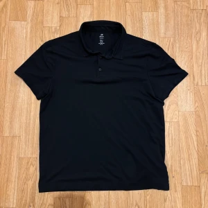 Svart pikétröja från H&M - Svart pikétröja från H&M med klassisk krage och två knappar framtill. T-shirten har korta ärmar och är gjord i mjuk bomull som känns skön mot huden. Enkel och stilren design som funkar till många olika looks. Det är storlek M men den hade passat S också