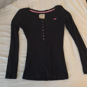 Svart långärmad tröja Hollister - Svart långärmad tröja från Hollister med knappar framtill och rosa logga på bröstet. Tröjan har en figurnära passform och är gjord i mjuk bomull, perfekt för en chill och stilren look.