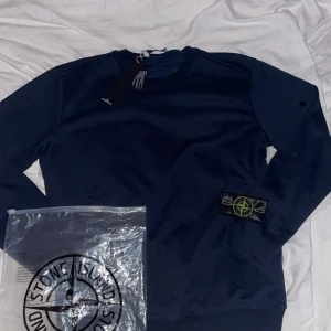 Mörkblå Stone Island sweatshirt - Mörkblå sweatshirt från Stone Island med klassisk rund hals och ikonisk patch på vänster ärm. Tröjan har ribbade muddar och är tillverkad i mjuk bomull. Perfekt för dig som gillar streetwear och vill ha en clean look.