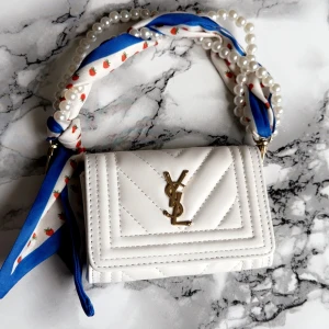 Vit quiltad YSL handväska med pärlhandtag - Superfin vit quiltad handväska från YSL med guldigt YSL-emblem framtill. Handtaget är dekorerat med pärlor och en blå/vit scarf med små jordgubbar. Perfekt accessoar för att lyfta din outfit! Inte äkta men jättebra qualitet. Mini väska 