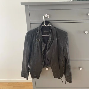 Svart skinnjacka från Hollies, stl 36 - Säljer en svart skinnjacka från Hollies i storlek 36. Jackan har coola detaljer som dragkedjor vid ärmslut och fickor, samt spännen i midjan och vid kragen. Snygg passform och klassisk bikerstil som lyfter vilken outfit som helst. Aldrig använd