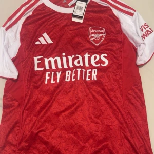 Arsenal fotbollströja  - Säljer en officiell Arsenal fotbollströja från Adidas i rött med vita detaljer och korta ärmar. Tröjan har ett subtilt mönster med Arsenal-loggor, broderad klubbmärke och Adidas-logga på bröstet samt Emirates Fly Better-tryck. Materialet är lätt och andas, perfekt för match eller träning.