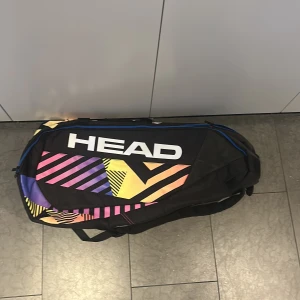 HEAD tennisväska med färgglada detaljer - Stor och rymlig tennisväska från HEAD med svart bas och färgglada grafiska mönster i rosa, lila, gul och orange. Väskan har dragkedja och bärremmar, perfekt för att ta med rack och utrustning till träningen.