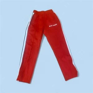 Röda mjukisbyxor från Palm Angels - Röda mjukisbyxor från Palm Angels med vita sidoränder och logga på vänster höft. Byxorna har elastisk midja och dragkedja vid benslut. Perfekta för en chill och sportig look.