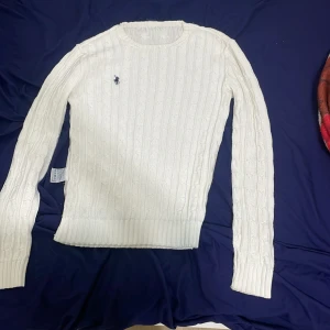 Vit kabelstickad tröja Ralph Lauren - Vit kabelstickad tröja från Ralph Lauren i 100% bomull. Klassisk rund halsringning, långärmad och med den ikoniska broderade loggan på bröstet. Perfekt för lager-på-lager och snygg till jeans eller kjol.