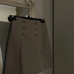 Beige/grå minikjol med knappar -  beige/grå kjol med eleganta guldfärgade knappar framtill. Storlek 32 men passar även 34. Knappt använd och i mycket fint skick