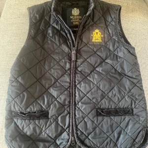 Svart quiltad väst från Morris - Quiltad svart väst från Morris med gul broderad logga på bröstet. Västen har dragkedja framtill, två fickor med manchesterdetaljer och en klassisk ståkrage. Perfekt för lager-på-lager och snygg till både jeans och chinos.