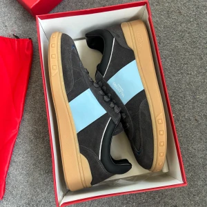 Valentino Garavani sneakers svart/blå - Snygga sneakers från Valentino Garavani i svart mocka med ljusblå detalj på sidan och beige gummisula. Klassisk rund tå, svarta skosnören och diskret branding på sidan och sulan. Perfekta för dig som vill ha en stilren men unik look.