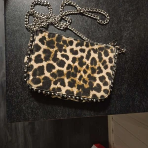 Leopardmönstrad axelväska med kedja - Cool axelväska med leopardmönster och silverfärgad kedja som axelrem. Väskan har dragkedja upptill och dekorativa nitar längs kanterna. Perfekt för dig som vill sticka ut med en trendig accessoar.