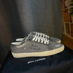 Grå Saint Laurent sneakers - Grå sneakers från Saint Laurent med vit sula och vita snören. Skorna har handskriven logga på sidan och är tillverkade i mocka med en klassisk låg profil. Perfekta för dig som gillar stilrena och exklusiva sneakers med en unik touch.