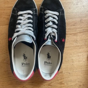 Svarta sneakers Polo Ralph Lauren - Svarta sneakers från Polo Ralph Lauren med vit sula, vita snören och rosa broderad logga på sidan. Ovandel i mocka och läder, insida i textil och sula i gummi. Snygga kontrastdetaljer med rosa häl och silverfärgad plös. Perfekta för en stilren och sportig look.