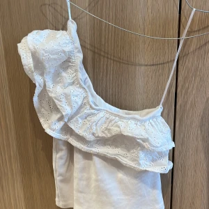 Vit oneshoulder topp med volang Gina Tricot - Supersöt vit oneshoulder topp från Gina Tricot med broderade volanger och smal axelrem. Toppen är i ribbad bomull och har en romantisk känsla med broderade blommor på volangen. Perfekt för sommarens varma dagar och snygg till både jeans och kjol. Är lite skrynklig men jag stryker innan paketering