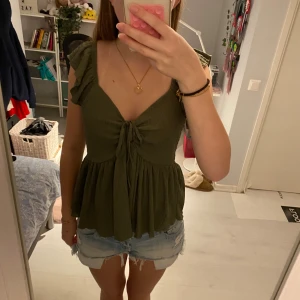 Olivgrön blus med knyt och volang - Supersöt olivgrön blus med volangärmar och knyt framtill från lager 157. Toppen har v-ringning, lätt rynkat tyg och en flowy passform som ger en avslappnad vibe. Perfekt till shorts eller jeans för en chill look.