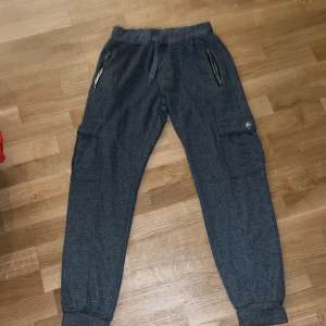 Grå mjukisbyxor med cargofickor - Mörkgrå mjukisbyxor med cargofickor på sidorna och dragkedjor vid fickorna framtill. Byxorna har resår och snörning i midjan samt muddar vid bensluten. Perfekta för en chill och avslappnad stil.