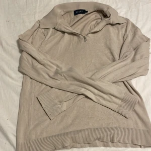 Beige stickad tröja med half zip från Riley - Beige stickad tröja från Riley i storlek S. Tröjan har en klassisk krage och half zip-dragkedja framtill. Långa ärmar och ribbade muddar vid ärmslut och nederkant ger en clean och stilren look. Perfekt för lager-på-lager under kyliga dagar.