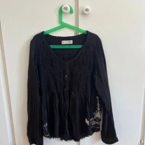 Svart blus från Odd Molly med broderi - Svart blus från Odd Molly med smockad överdel, knappar framtill och broderade detaljer i vitt och beige längs nederkanten. Långärmad modell i mjuk bomull med avslappnad passform och rundad halsringning. 