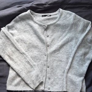 Ljusgrå stickad kofta från 157 - Mysig ljusgrå stickad kofta från 157 med rund halsringning och knappar framtill. Koftan har lång ärm och ribbade muddar vid ärmslut och nederkant. Perfekt för lager-på-lager och enkel att matcha med allt.
