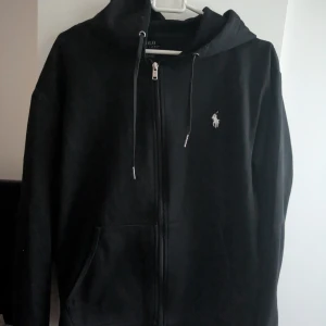 Svart hoodie från Polo Ralph Lauren - Svart hoodie från Polo Ralph Lauren med dragkedja framtill, snörning i huvan och klassisk broderad logga på bröstet. Tröjan är aldrig använd
