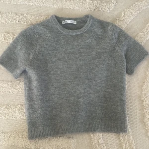 Grå stickad kortärmad tröja - Mysig grå stickad tröja från Zara i kortärmad modell. Tröjan har rund halsringning och ribbade kanter vid ärmslut och nederkant. 