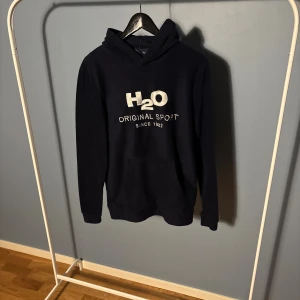 Mörkblå fleece hoodie från H2O - Mysig mörkblå hoodie från H2O med broderad logga och text på bröstet. Tillverkad i mjuk fleece, har huva och långa ärmar. Perfekt för chill dagar eller när du vill ha något extra bekvämt på dig.