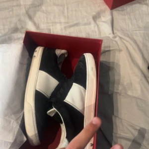 Valentino Garavani svartvita sneakers - Svartvita Valentino Garavani sneakers med klassisk låg siluett. Skorna har svarta mockadetaljer, vita skinnpaneler och ikoniska nitar längs sulan. Snygg kontrast mellan materialen och en stilren look som sticker ut.