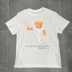 Ralph lauren t shirt  - Vit t-shirt från Ralph Lauren med ikoniskt Polo Bear-tryck där nallen håller en tennisracket. Klassisk rund hals och korta ärmar. Trycket är detaljerat och t-shirten har en clean vibe som passar dig som gillar sportiga och stilrena plagg.