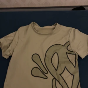 T shirt från syna world - Snygg grön t-shirt med stort grafiskt tryck i svart på både fram- och baksidan. T-shirten har rund hals och korta ärmar, perfekt för dig som gillar streetstyle och unika prints. Materialet känns mjukt och lätt, troligen bomull eller polyester.