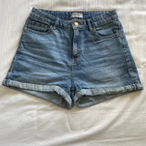 Jeansshorts - Snygga blå jeansshorts från Lindex med klassisk femficksdesign och uppvikta benslut. Shortsen har hög midja och stängs med knapp och dragkedja. Perfekta för varma dagar och enkla att matcha med allt i garderoben.