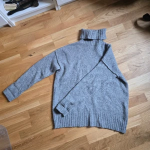 Grå stickad polotröja - Mysig grå stickad polotröja med hög, ribbad krage och långa ärmar. Tröjan har en relaxed passform och breda ribbade muddar vid ärmslut och nederkant. Strl S