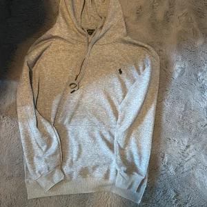 Grå hoodie från Polo Ralph Lauren - Snygg ljusgrå hoodie från Polo Ralph Lauren med klassisk huva och snörning. Tröjan har lång ärm, ribbade muddar och den ikoniska lilla broderade loggan på bröstet. Perfekt för en chill och stilren look.