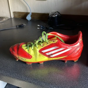 Adidas F50 röda och gula fotbollsskor - Säljer ett par Adidas F50 fotbollsskor i rött och gult med gröna snören och vita ränder på sidorna. Skorna har plastsula med fasta dobbar och är tillverkade i syntetmaterial. Perfekta för dig som vill sticka ut på planen!