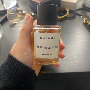 Essnce Irresistible Enigma parfym - En stilren parfymflaska från Essnce med doften Irresistible Enigma. Flaskan är cylindrisk i genomskinligt glas med svart lock och vit etikett. Innehållet är gyllengult och flaskan har en modern, minimalistisk design. Eau de Parfum.
