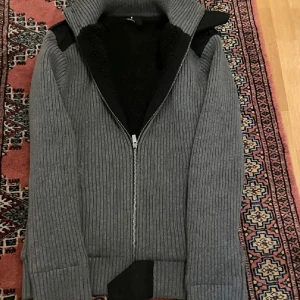 Grå Cardigan från Selected - Säljer en grå ribbstickad kofta från Selected med hel dragkedja och hög krage. Insidan är fodrad med svart teddyfoder för extra värme. Snygga svarta detaljer på axlarna och en stilren look som passar perfekt till kyliga dagar.