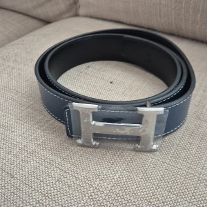 H-bälte  - Snyggt bälte i blått och svart skinn med ikoniskt,H-spänne i silverfärgad metall. Bältet har stilrena, kontrastsömmar och klassisk design som lyfter, vilken outfit som helst. Strl 110cm