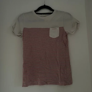 Randig t-shirt från Newbie, vit/röd - Säljer en söt t-shirt från Newbie i storlek 122/128. Den har vit överdel och röda smala ränder nedtill, samt en liten bröstficka med Newbie-logga. Rund hals och korta ärmar, tillverkad i mjuk bomull. 